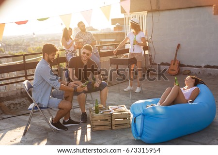 Group of young friends having fun at rooftop party, making barbecue, drinking beer and enjoying hot summer days. Focus on the man in black T-shirt