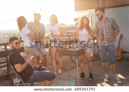 Group of young friends having fun at rooftop party, making barbecue, drinking beer and enjoying hot summer days. Focus on the couple next to the barbecue