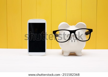 Piggy bank wearing a glasses with a cellphone over yellow wooden wall