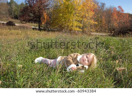Adorable little blond girl laying in the grass outside with trees changing colors in autumn