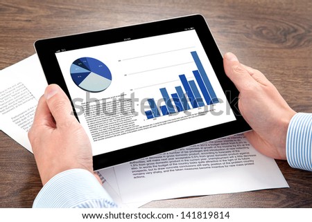 Businessman Holding A Tablet Computer With Graphics On A Screen On The Table With Documents