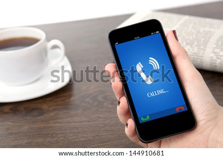 Businesswoman Hand Holding A Phone With Ringing Phone Receiver On A Screen Against The Background Of A Table In The Office
