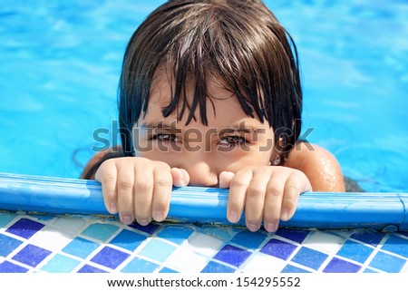 Wet little girl with beautiful eyes peeking out of the pool with blue water