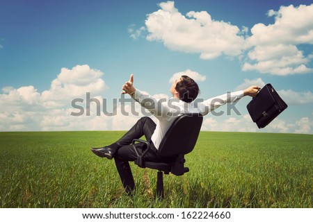 businessman in a suit in a green field with a blue sky sitting on an office chair and waving his arms