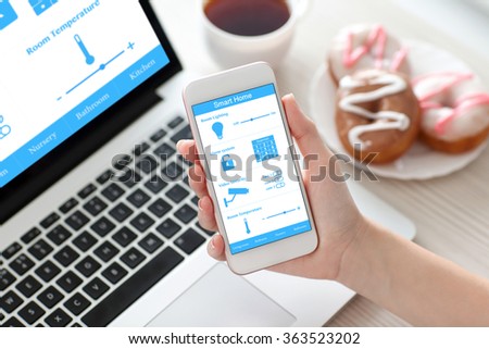 female hand holding white phone with program smart home on the screen against the background of the computer