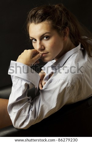Portrait of young beautiful girl in men\'s shirt sitting in black chair on black background