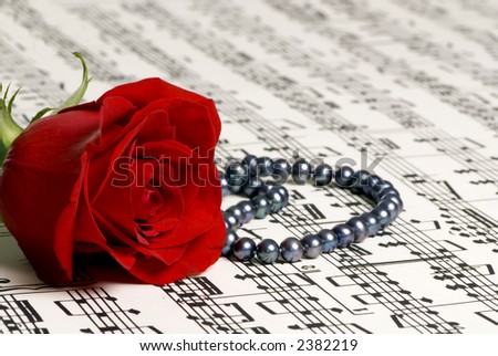 A single red rose lays on top of a complicated score of music. A gift of black pearls sits next to the rose.