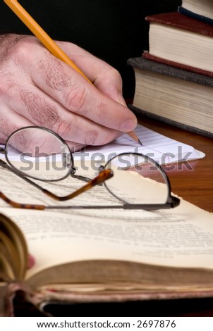 A person works on a paper with open books and glasses on a table
