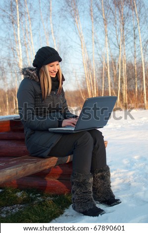 Smiling beautiful young woman watching laptop sitting on a bench in winter surrounded by snow