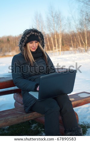 Smiling beautiful young woman watching laptop sitting on a bench in winter surrounded by snow