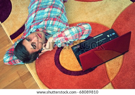 top view of handsome young man lying on back on the carpet floor with laptop while listening to phone