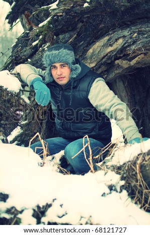 Fashion portrait of a crouching handsome young man enjoying himself in winter in front of big old tree trunk