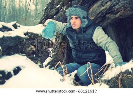 Fashion portrait of a crouching handsome young man looking at something in winter in front of big old tree trunk