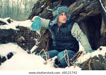 Fashion portrait of a crouching handsome young man looking at something in winter in front of big old tree trunk