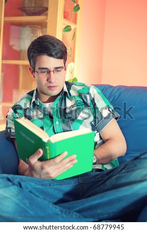Portrait of an attractive young man with glasses reading the book on couch in his living room
