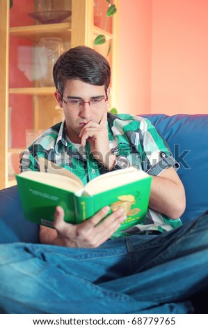 A thoughtful handsome student with glasses relaxing reading a book on a couch in a living room