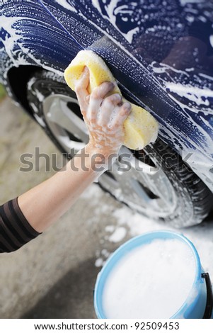Man washing blue car with a yellow sponge and a bucket of soapy water.
