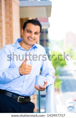 Portrait of good looking businessman giving you a thumbs up, enjoying a drink, standing on his balcony - Isolated on a city background