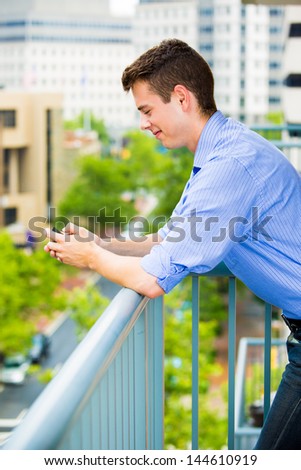 Portrait of handsome guy using his cell phone, texting, web, while relaxing on outside balcony, isolated on background of buildings and trees
