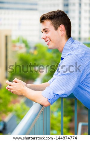 Portrait of handsome guy using his cell phone, texting, or browsing web, while relaxing on outside balcony, isolated on background of buildings and trees