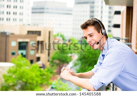 Closeup portrait of customer service representative or call center agent or support staff or operator with headset on outside balcony, isolated on outside background with trees and city buildings