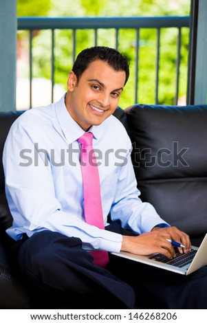 Closeup portrait of young, handsome student or businessman or lawyer, working on laptop, on a black couch at his home or office, isolated on city background with trees