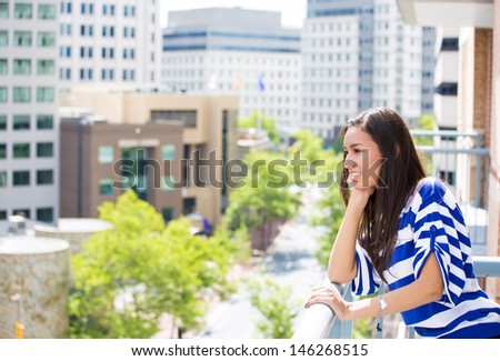 A portrait of a beautiful young female relaxing on a balcony on a sunny summer day, in her new apartment, background of a city scenery and green trees. Urban lifestyle.