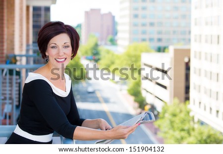 A portrait of a beautiful smiling female, businesswoman, relaxing on a balcony on a sunny summer day, reading a newspaper, on a background of a city buildings and green trees. Urban student lifestyle