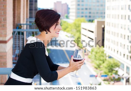 A portrait of a beautiful female, businesswoman relaxing on a balcony on a sunny summer day,drinking red wine, isolated on a background of a city buildings and green trees. Urban corporate lifestyle