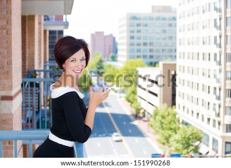 A portrait of a beautiful female, businesswoman relaxing on a balcony on a sunny summer day,drinking tea or coffee, isolated on a background of city buildings, green trees. Urban corporate lifestyle