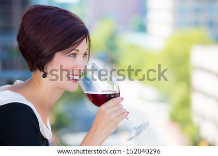 A close-up portrait of a beautiful female, businesswoman relaxing on a balcony on a sunny summer day,drinking red wine, isolated on a background of city buildings and green trees. Urban lifestyle