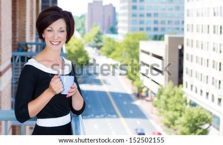 A portrait of a beautiful female, businesswoman relaxing on a balcony on a sunny summer day,drinking tea or coffee, isolated on a background of city buildings, green trees. Urban corporate lifestyle