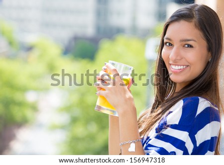A Portrait Of A Beautiful Young Female Relaxing On A Balcony On A Sunny Day, Drinking Orange Juice, In Her New Apartment, On A Background Of A City Scenery And Green Trees. Urban Student Lifestyle