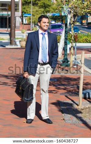 A whole body portrait of a young smiling, happy, successful business man, executive walking outside down the street holding a briefcase isolated on a background of a city