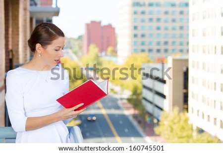Closeup portrait of beautiful, young, happy, relaxed woman enjoying a weekend on sunny day, on balcony of her apartment, reading a book, isolated on city background. Urban life style of businesswoman