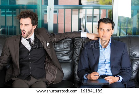 Closeup portrait of two businessman sitting on black couch waiting to talk someone important or an office appointment or interview.  One guy is bored.  Other is pissed off. Emotions and communication