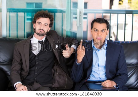 Closeup portrait of two co-workers, businessmen, corporate or government employees sitting on black couch, one giving middle finger, other thumbs up sign isolated on city background. World polarity