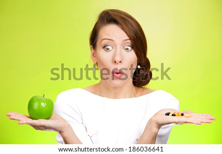 Closeup portrait young confused, puzzled woman holding green fresh apple in one hand, pill, vitamins in another, trying to decide which choice is the best one. Face expressions, emotions, health care.