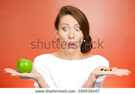 Closeup portrait young confused, puzzled woman holding green fresh apple in one hand, pill, vitamins in another, trying to decide which choice is the best one. Face expressions, emotions, health care.