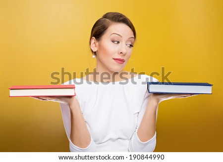 Closeup portrait, young business woman confused student holding red, blue books in hands, deciding which one to choose, way to go isolated yellow background. Emotion, expressions, reaction, perception