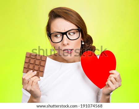 Closeup portrait, headshot beautiful, young nerd woman with black glasses holding heart craving square milk chocolate isolated green background. Food diet option dilemma. Sweet temptation. Expression