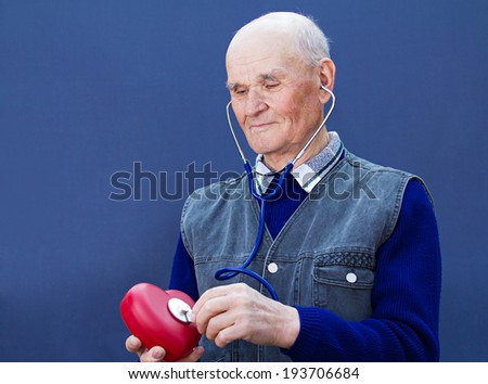 Closeup portrait mature, senior, elderly man, grandfather with stethoscope listening to red heart isolated blue background. Obamacare, universal health plan. Cardiovascular disease stress, free life