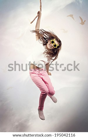 Portrait happy, smiling little girl in sunglasses hanging on rope with one hand, somewhere in space, sky, isolated clouds background. Human life fate, challenges, destiny, luck, karma, chance concept