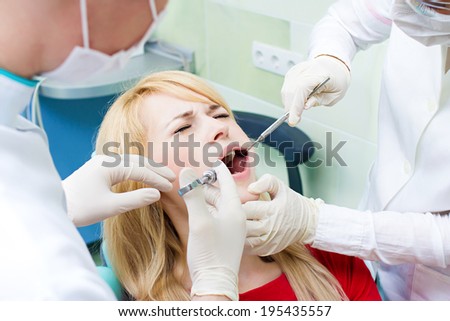 Closeup portrait young female, woman patient sitting in dentist chair, office with wide open mouth getting painful anesthetic injection done by doctor, his assistant isolated background clinic office