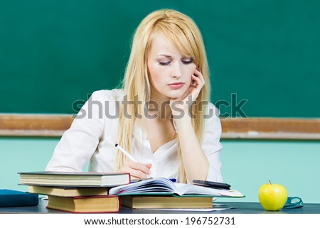 Closeup portrait young blonde woman, student sitting at desk studying in library, class room, isolated background green chalkboard. Facial expression, education, college, university life style concept
