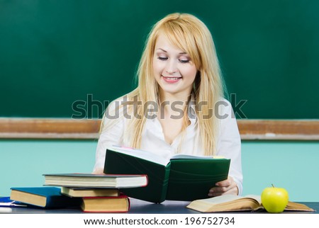 Closeup portrait young blonde woman, smiling student sitting at desk studying in library class room isolated background chalkboard. Facial expression, education, college, university life style concept