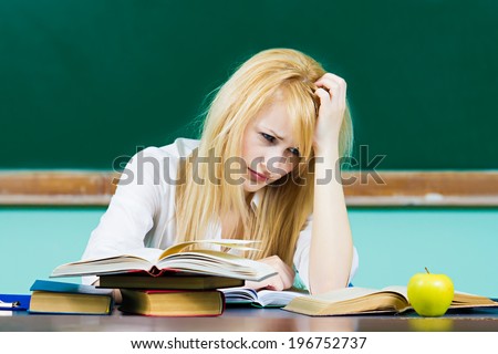 Closeup portrait young worried woman, confused student sitting at desk studying in library class room isolated background chalkboard. Facial expression, education college university life style concept