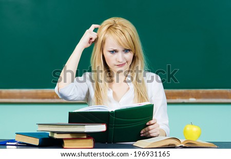Closeup portrait young blonde woman, confused student scratching her heard sitting at desk in classroom thinking hard, reading book puzzled isolated background with chalkboard. Human facial expression