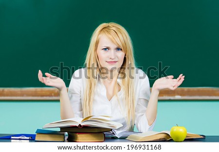 Closeup portrait blonde woman, confused student shrugging her shoulders has no answer, sitting at desk in classroom, thinking, puzzled isolated background chalkboard. Human emotions, facial expression