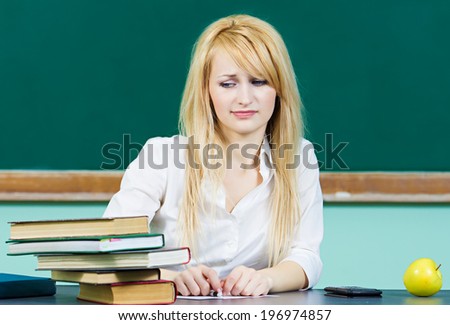 Closeup portrait hopeless skeptical overwhelmed student sitting at desk in front pile book studying preparing for finals isolated chalkboard background. Education college life concept. Face expression